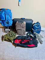 All six items arranged on tiled floor against a beige wall: includes varying types of luggage bags and a camouflage military vest.