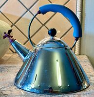 Front and side view of polished stainless steel tea kettle with blue handle and bird whistler on spout, sitting on granite countertop.