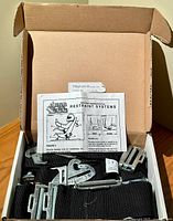 Opened box showing uninstalled Safety Solutions restraint system with black straps and multiple metal fitting components, including buckles and mounting hardware, with installation instructions visible.
