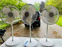 Front view of three white oscillating floor fans with protective grilles and three blades each, displayed on a table outdoors.