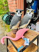 Photo showing two gray owl ornaments and one pink flamingo ornament on a wooden surface outside near brick wall and garden hose.