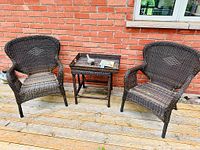 Wide shot of two brown PVC resin woven wicker armchairs and matching side table on wooden deck against brick wall.