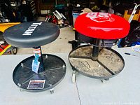 Two mechanic stools side by side, showing black and red vinyl seats, circular metal bases with tool trays and caster wheels.