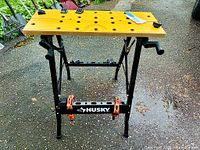 Front view showing the wooden top with holes, metal legs, and orange clamps attached.