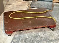 Primitive wooden dolly platform with yellow rope on a concrete floor.