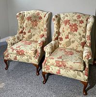 Photo shows two Queen Anne style wingback chairs with floral upholstery, placed side by side on a carpet. Both have wooden cabriole legs, one chair has a dark spot near the top of the backrest.