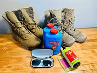 Both pairs of tan Red Head hunting boots side by side with two sealed Gold Bond powder containers, a lint roller, and sunglasses hard case on wood surface.