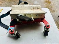 Top-front angled view showing the white support platform with scratches, black securing strap, and red metal base with caster wheels.