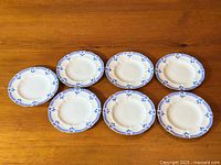 Set of 7 Royal Crown Derby 'Grenville' bread and butter plates arranged on wooden table showing overall pattern and quantity.