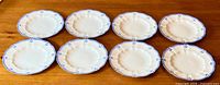 Eight white fine bone china dinner plates arranged in a group on a wooden surface, showing the front with blue floral border and scalloped edge design.