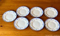 Seven white fine bone china salad or side plates with blue floral Grenville pattern edges arranged on a wooden surface