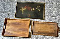 Overhead photo showing three serving trays, one black lacquered with butterflies and flowers, two natural wood with handles and rope detail