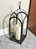 Tall black metal open frame lantern with arched design, featuring a clear glass dome inside and a white candle placed within. The lantern sits on a tiled floor next to a wooden door.