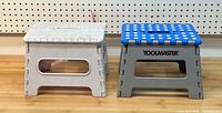 Photo showing front view of two foldable two-step stools side by side, one white and one grey with blue top and TOOLMASTER logo.