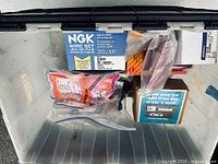 Top-down photo showing assorted car parts in a plastic bin with visible NGK wire set, Stewart Warner bolt, and other components.