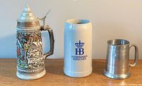 Three vintage beer steins displayed side by side: one with pewter lid and colorful painted ceramic, one with blue logo Hofbrauhaus München ceramic stein, one small metal stein.