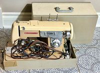 Vintage Domestic Tessler sewing machine in beige with cords on base, shown outside carrying case on floor.