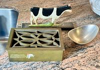 Metal cow door stop, brass vent cover, and Nambe modern dish arranged on countertop.