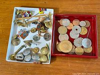 Overview of two trays with assorted military buttons, pins, coins and cufflinks displayed on a wooden surface.