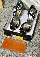 Photo showing floral patterned ladies heels shoes in box with visible black straps on open-toe design, leather wallet on carpet.