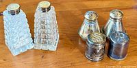 Pair of clear glass salt and pepper shakers with silver tops alongside two pairs of smaller silver and silver-plated shakers on wooden surface.