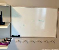 Photo shows the wall-mounted whiteboard installed on wall beneath a shelf, white frame with visible marker residue, black Expo dry erase board eraser resting on bottom ledge, several boxes of markers and accessories on the shelf below.