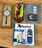 All items arranged on a wood surface, showing the multimeter with attached test leads, dial indicator in box, AcuAngle angle level in sealed package, and Kestrel pocket wind meter box.