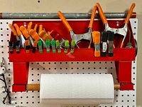 Full front view of the red metal wall-mounted towel rack with assorted spring clamps, small clips and a paper towel roll on wooden dowel holder.