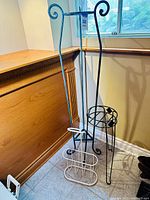 Photo of two wrought iron plant stands and a white wire umbrella holder arranged in corner by window.