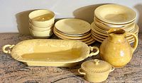 Overview of large pile of yellow stoneware dishes including varied sizes of plates, bowls, a rectangular serving platter with vine decoration, a covered casserole, and a pitcher.