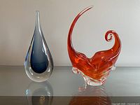 Photo of two glass sculptures on glass surface against white wall: blue teardrop shape and red curling design.