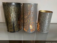 Three metal candle holders with tree silhouette cutout design, varying heights, silver exterior and gold interior, displayed on glass surface.