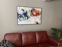 Abstract multicolor painting print hanging on wall above red leather sofa, showing full artwork and frame.