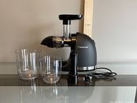 Black Amzchef slow juicer with two clear juice containers on a table against a plain wall.