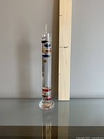 Full view of tall glass Galileo thermometer standing next to wooden ruler showing approximate 15 inch height.