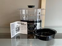 Full view of Hamilton Beach digital steamer unit with instruction manual and extra black tray placed next to it.