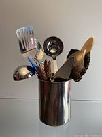 Photo shows a variety of kitchen utensils including stainless steel slotted spatulas, ladle, wooden spoon, silicone spatulas and basting brush, stored in a stainless steel holder.