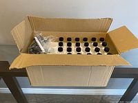 Photo of large open cardboard box showing 50 clear glass bottles with black screw caps inside, with some accessories on top in plastic bags.