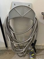 Coiled silver metal garden hose hanging on wall-mounted gray hose holder with spray nozzle attached, showing overall setup and condition.