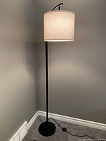 Full view of black metal floor lamp with white fabric shade placed in corner against grey walls and white baseboard.