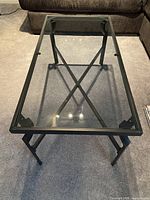 Top view of rectangular coffee table showing clear glass tabletop, black metal frame with crossbar reinforcement beneath.