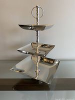 Front view of the assembled modern silver 3-tier metal serving tray with square plates of different sizes and circular handle on top.