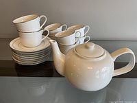 Eight white porcelain teacups with silver trim stacked on top of matching saucers next to a white porcelain teapot.