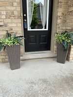 Pair of tall brown synthetic wicker outdoor planters with artificial fern leaves and small yellow flowers placed on either side of an exterior front door.