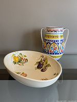 Photo showing the white oval pottery bowl decorated with colorful vegetable illustrations alongside the tall vase with vibrant multicolored patterns and two handles.