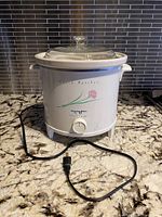 Front view of Proctor-Silex slow cooker with power cord placed on granite countertop, showing floral design and control dial.
