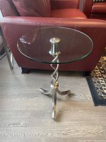 Full view of the side table showing the glass top and the swirled metal base with curved feet, positioned on a wooden floor beside a leather sofa.