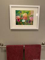 Framed floral watercolor artwork showing pink and purple irises in a white frame, hung on a neutral wall above red towels