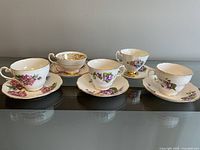 Five vintage fine bone china teacup and saucer sets arranged on a glass surface, showing floral patterns including pink roses, purple violets, and other flowers, with gold trim.