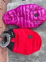 Photo shows two Canada Pooch dog jackets on pavement, one pink and one red with black and faux fur hood